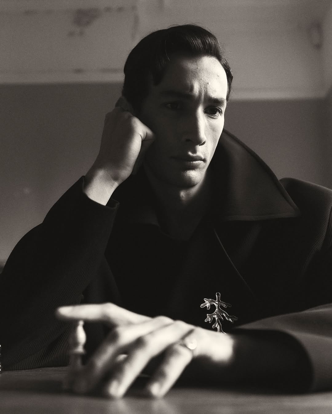 Portrait of a man with dark hair, resting his head on his hand while sitting at a table with a chess piece in front of him.