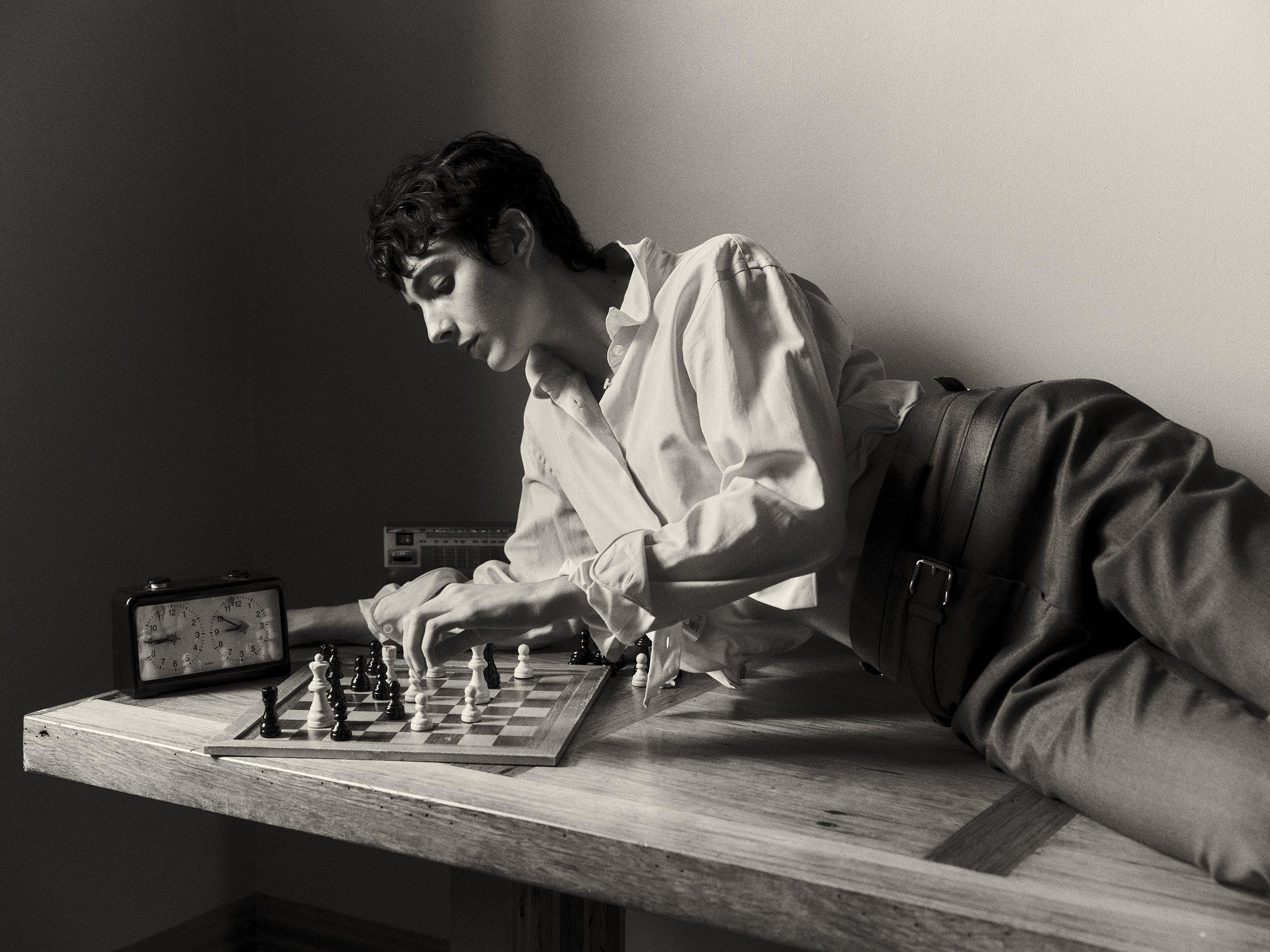 Monochrome portrait of a young adult playing chess at a table, featuring a vintage clock, against a neutral background.