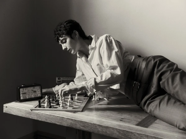 Monochrome portrait of a young adult playing chess at a table, featuring a vintage clock, against a neutral background.