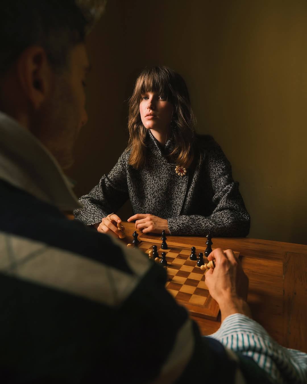 Woman in grey tweed jacket plays chess with a man in an indoor setting; focus on chess pieces and board.