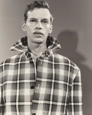 Portrait of a man wearing a plaid button-down shirt with a large popped collar against a grey background.
