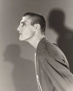 Profile portrait of a young man wearing a zipped jacket against a plain grey background with a strong shadow cast behind him.
