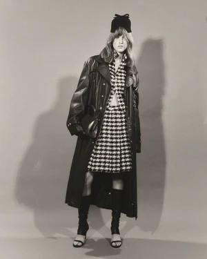 Model wearing a black leather trench coat, houndstooth skirt suit, and bow hat holding a handbag against a plain backdrop.