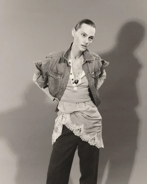 Model wearing a denim vest, layered top with lace trim, and dark trousers against a grey seamless studio backdrop with a harsh shadow.