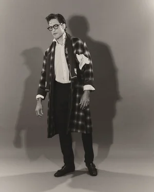 Man wearing thick-rimmed glasses, a white shirt, dark trousers, and a plaid robe standing against a plain backdrop.