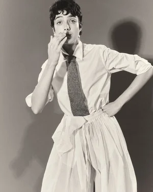 Model in white collared shirt, dark tie, and light skirt against a plain studio background with cast shadow.