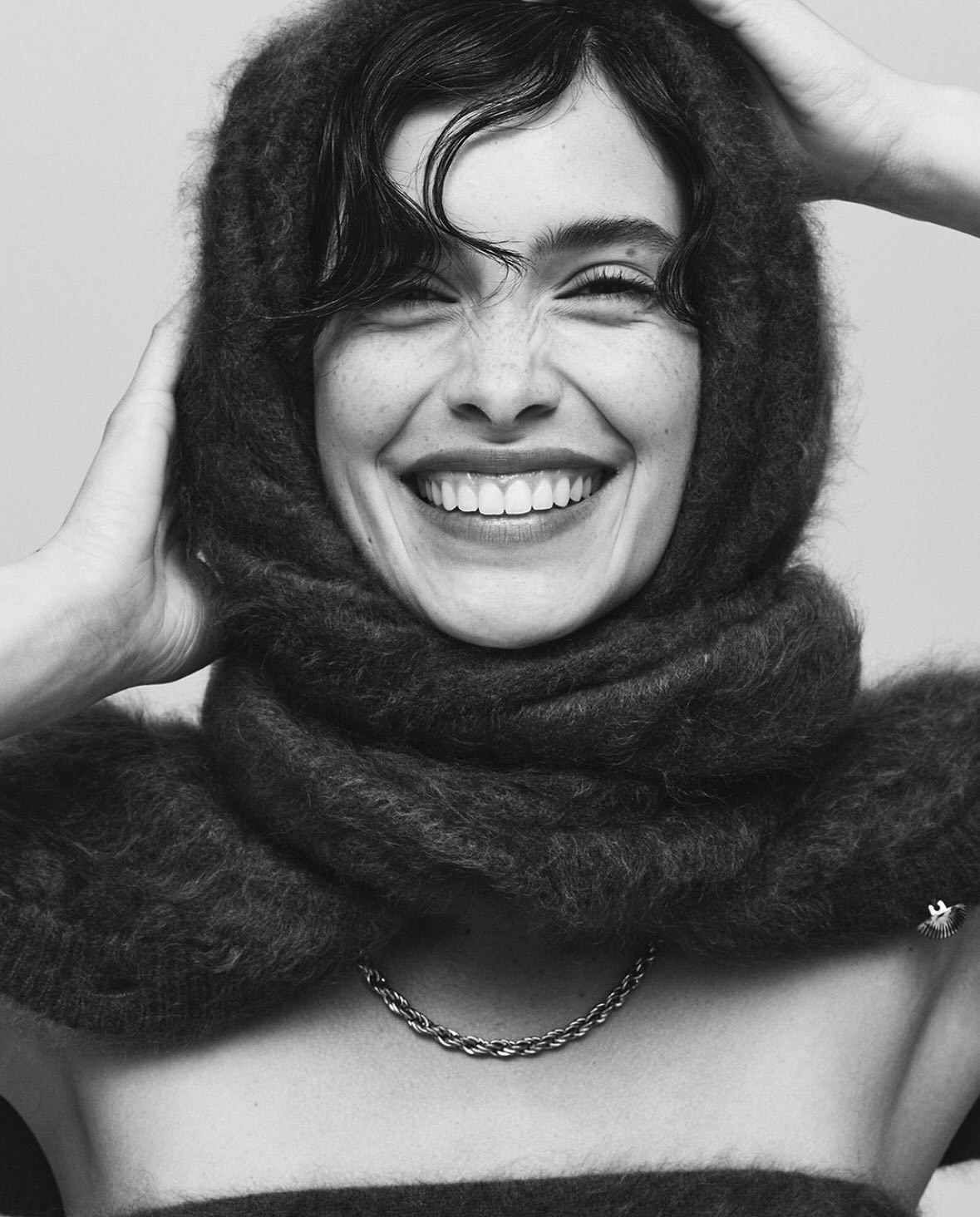 Black and white portrait of a smiling woman with a dark, fuzzy scarf around her head and shoulders. She also wears a chain necklace.