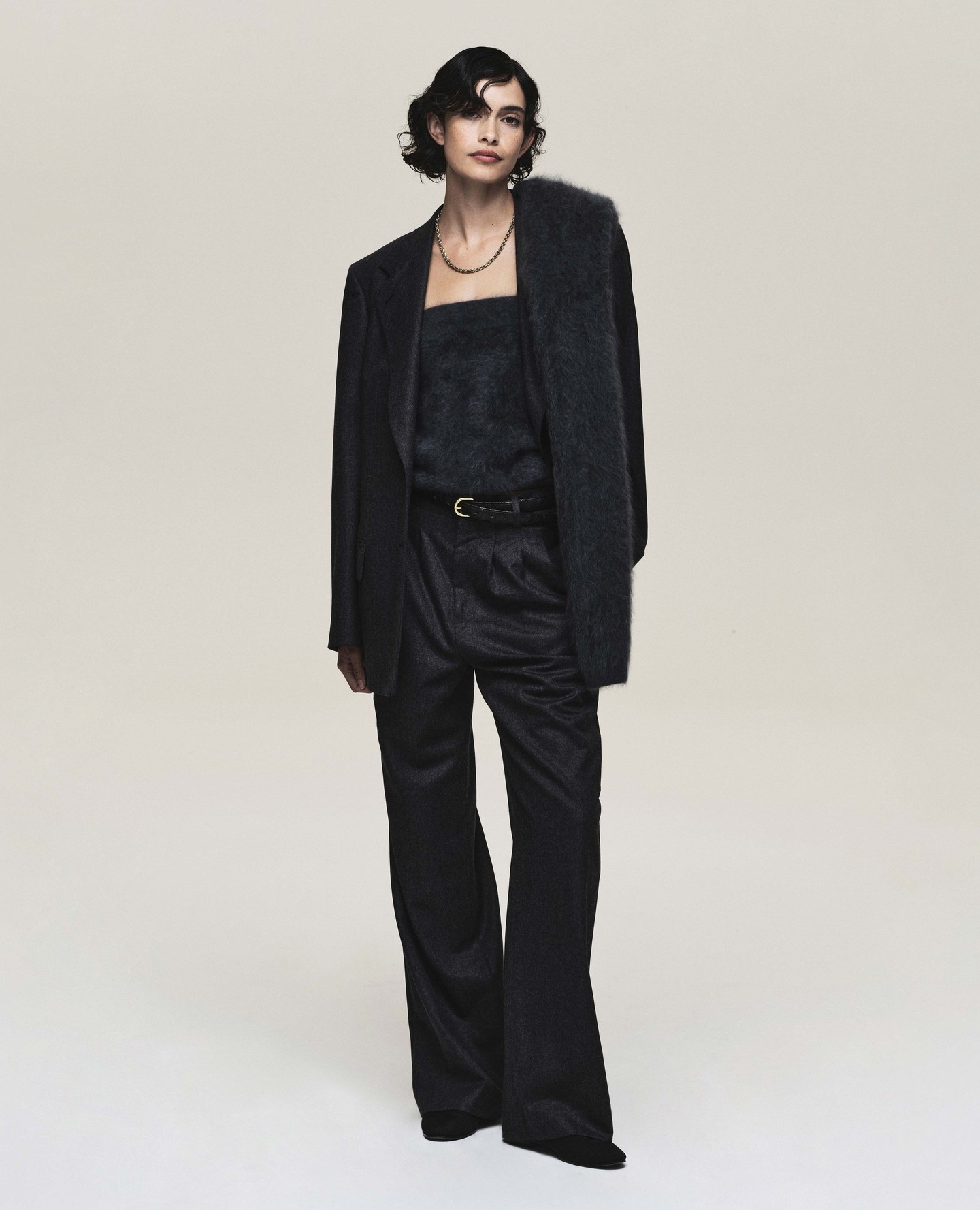 A woman in a dark blazer, top, and slacks poses in a full shot against a light backdrop, showcasing a fashion-forward, elegant style.