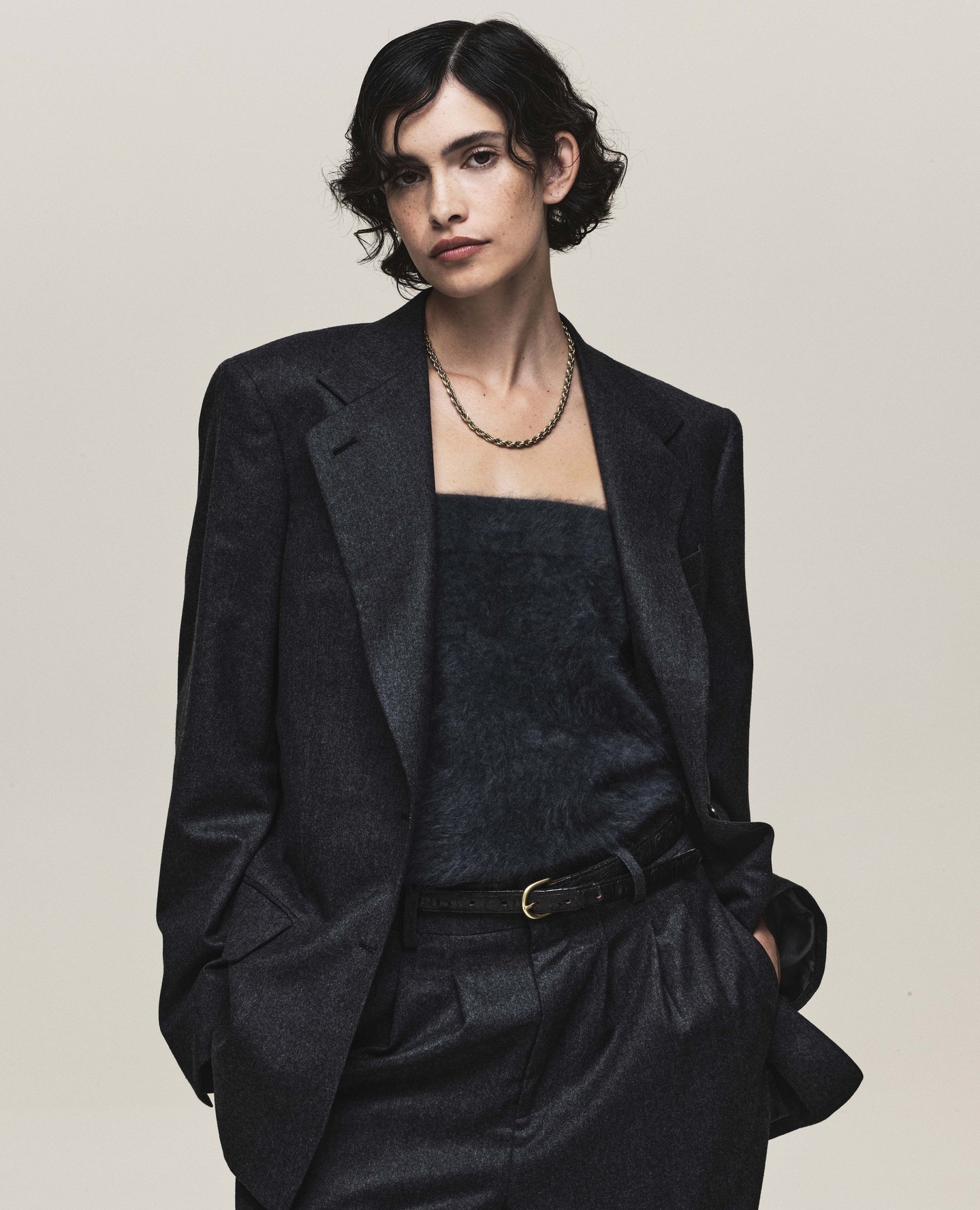 A woman in a dark-grey wool suit and tank top poses indoors against a white backdrop in a medium shot with soft, natural lighting.