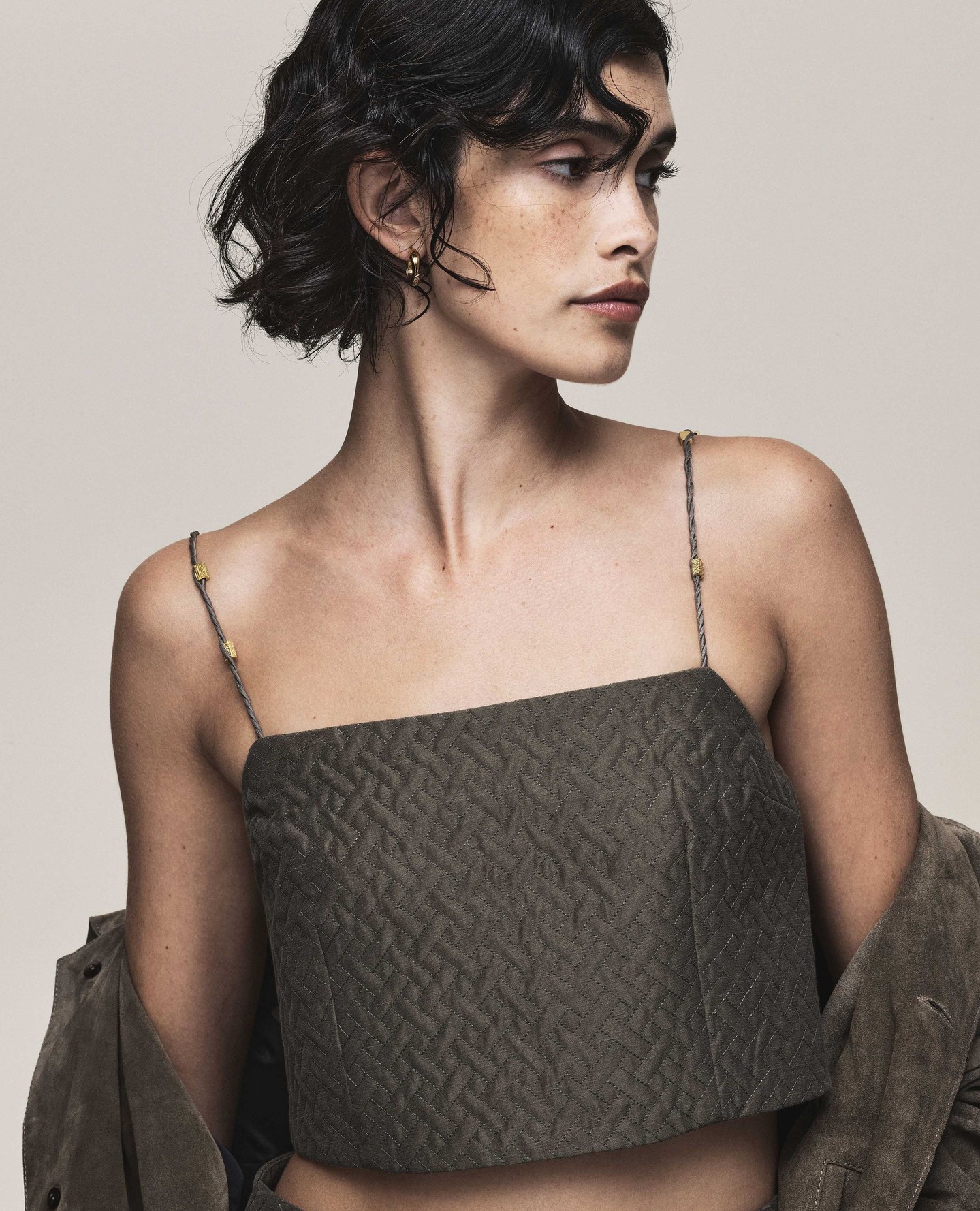 A brunette adult woman poses indoors wearing a patterned top with thin straps, showcasing an olive green jacket off the shoulder.