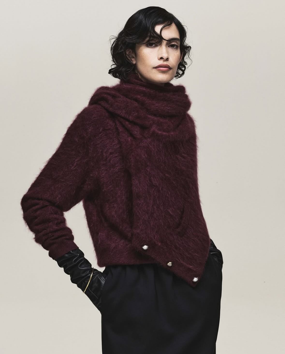 A brunette fashion model poses in a maroon mohair-style hooded sweater with black trousers and gloves against a neutral studio backdrop.