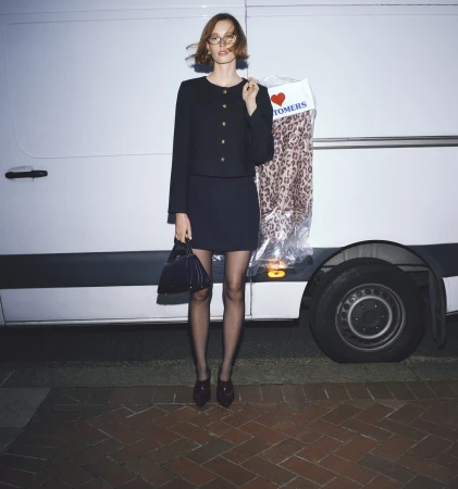 A woman in a dark suit poses with a garment bag and purse in front of a white van with brick flooring.