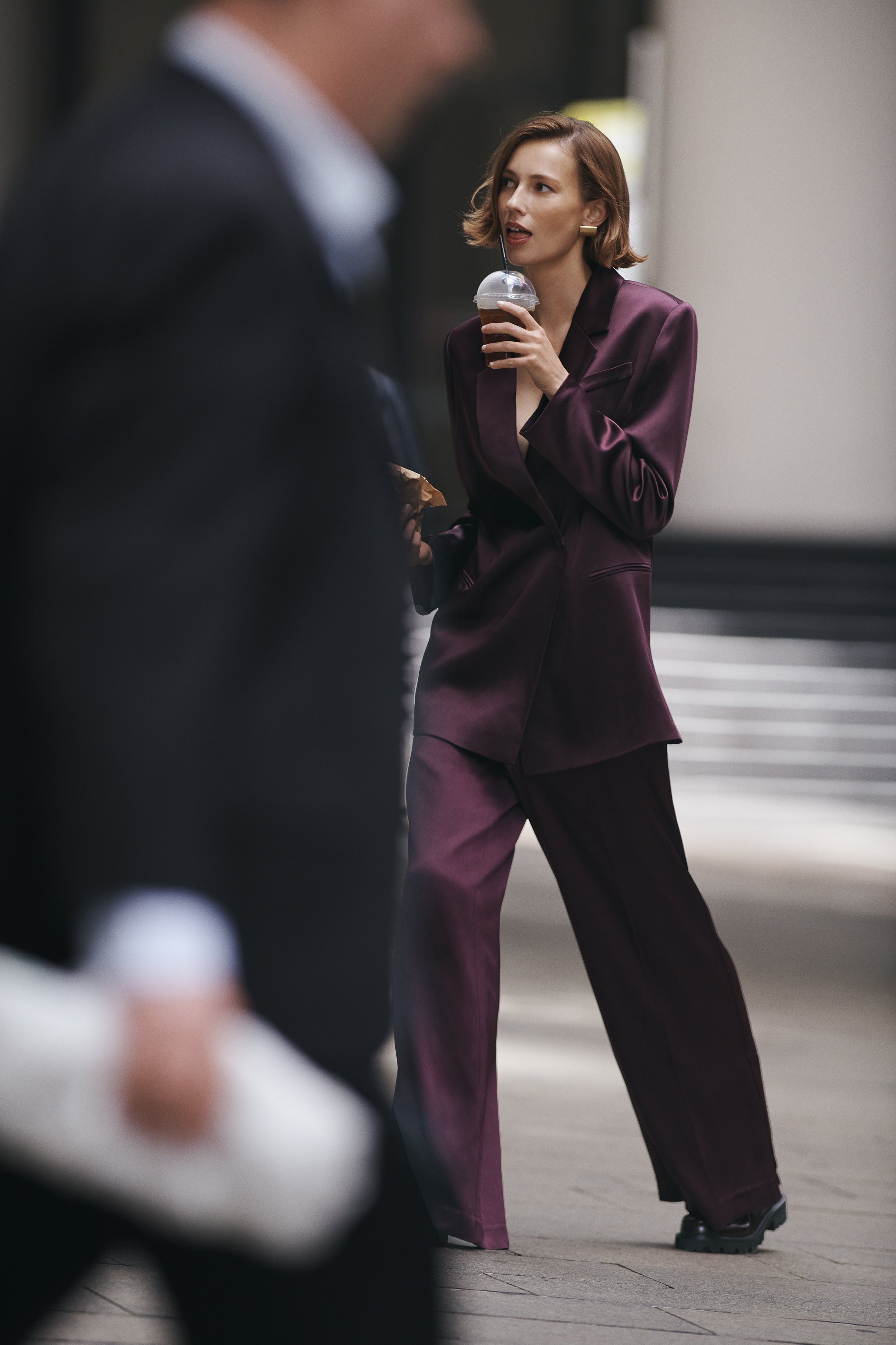 Stylish woman in plum suit walks outdoors with coffee, passerby in suit visible in foreground, capturing a candid street-style scene.