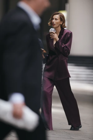 Stylish woman in plum suit walks outdoors with coffee, passerby in suit visible in foreground, capturing a candid street-style scene.