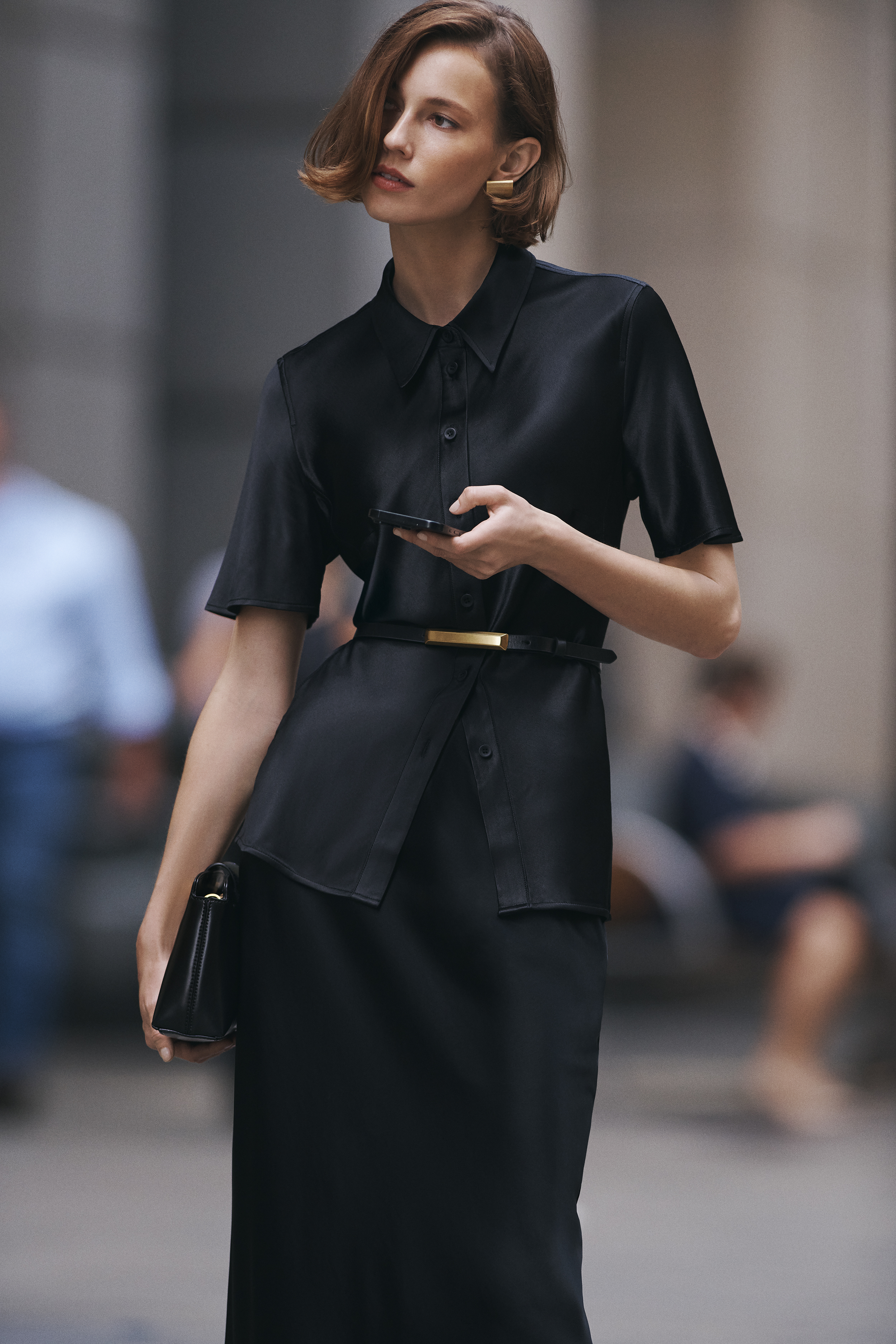 Woman in black shirt dress and gold earrings holding a phone, standing outdoors with blurred people in the background on a city street.