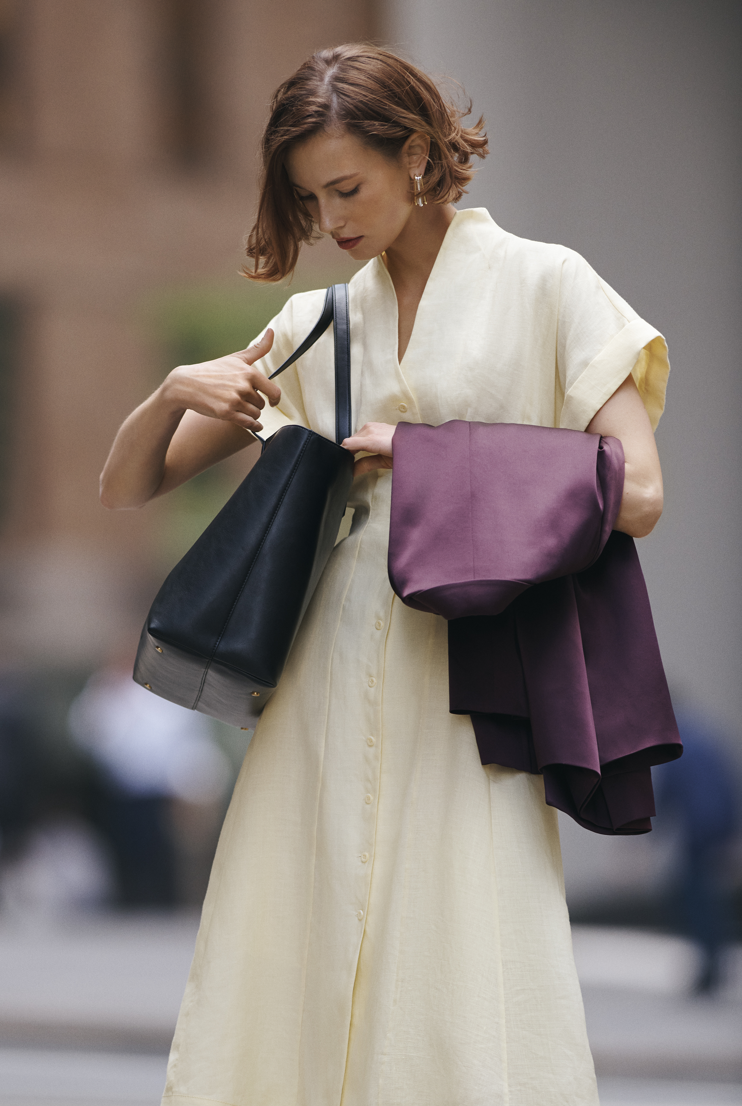 Adult woman in a light yellow dress outdoors looking into her black tote bag. She also carries a purple shawl over her shoulder.