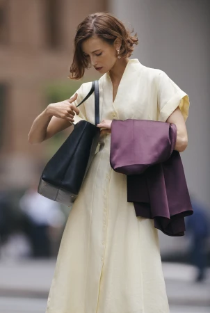 Adult woman in a light yellow dress outdoors looking into her black tote bag. She also carries a purple shawl over her shoulder.