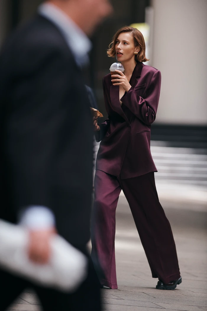 Woman in a plum satin pantsuit holding an iced drink on an urban street with a blurred figure in the foreground.