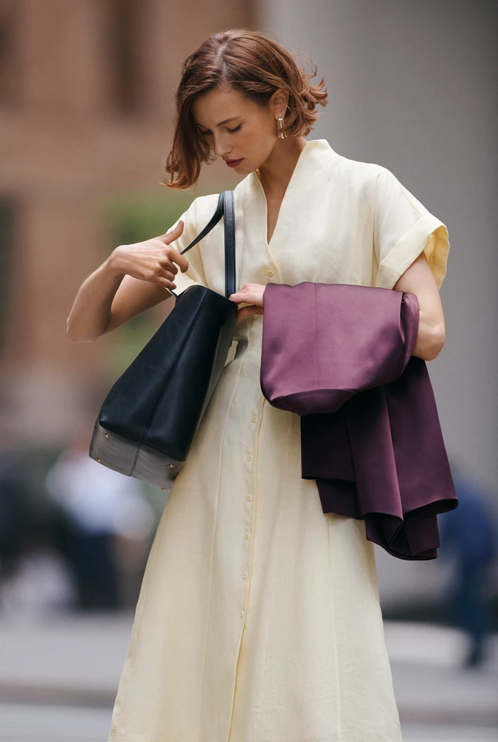 Woman in cream dress holding a dark handbag and a plum jacket on an urban street.
