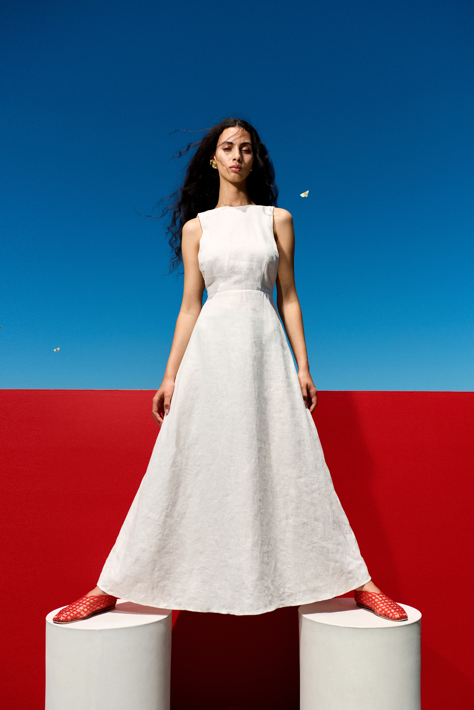 A woman stands on two white columns in a sleeveless white gown with a blue and red background, posing for a fashion shot.