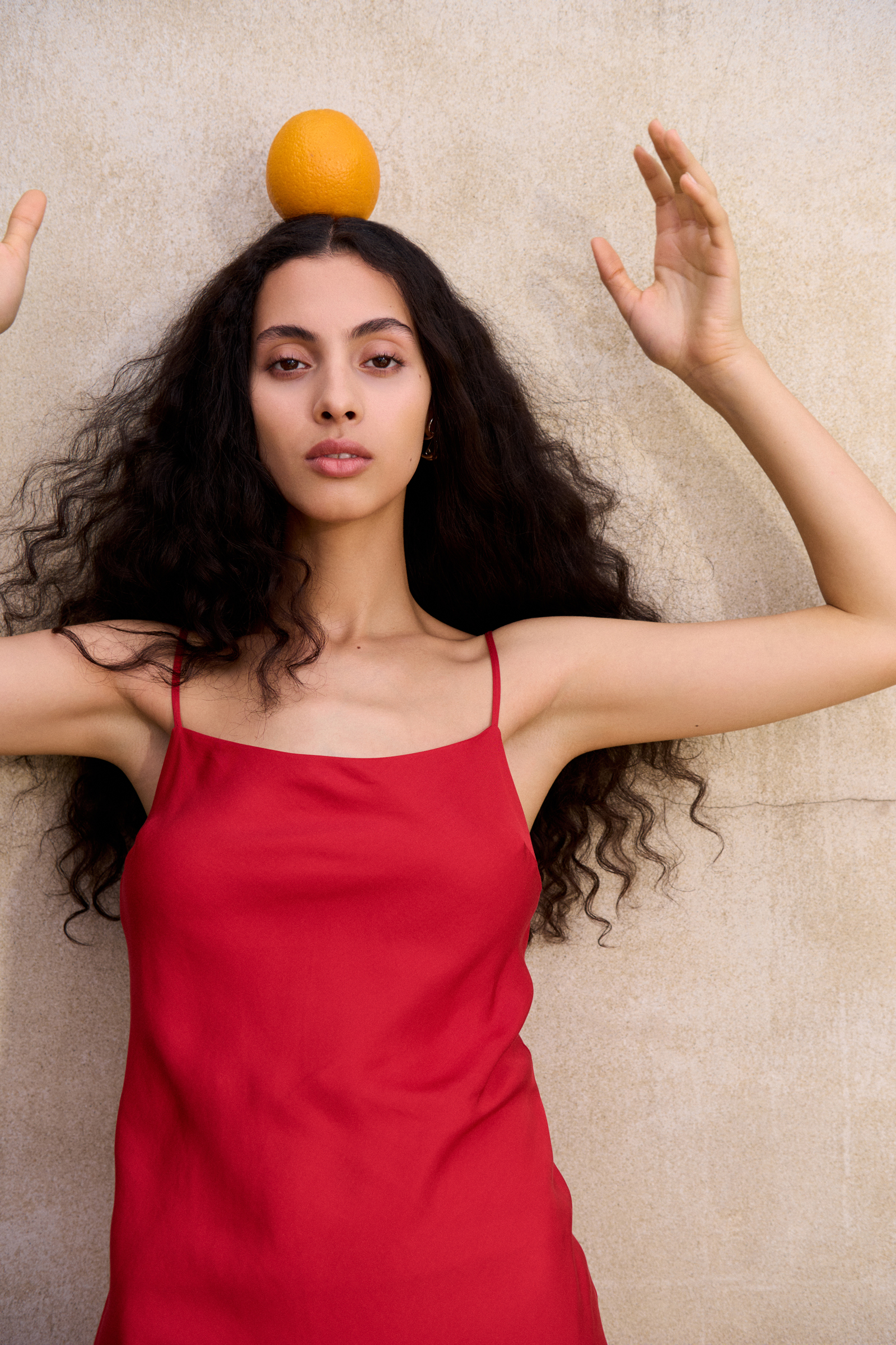 Woman in red dress with orange on her head poses against a neutral wall, arms raised, looking at the camera with a serious expression.
