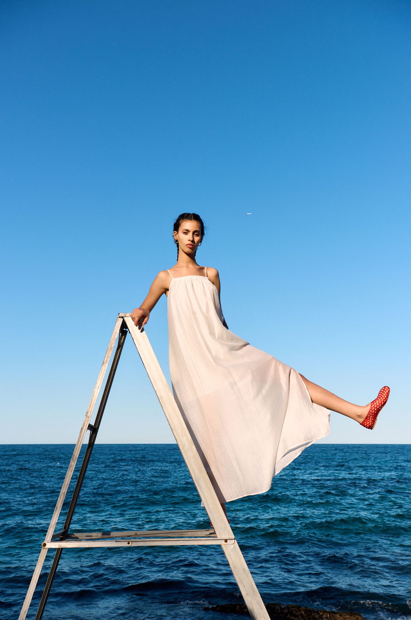 Woman in a sundress climbs a silver ladder against a blue sky and ocean backdrop, creating a serene and stylish summer scene.