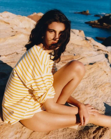 Woman in a yellow-striped shirt poses on rocks by the ocean. Close-up shot of a young adult female near the sea in daylight.