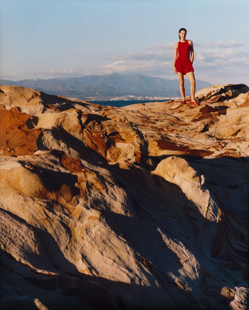 A woman in a red dress stands atop a rocky hill with mountains and water visible in the background under a clear blue sky at sunset.