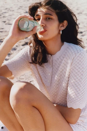 A woman with dark hair sits on a beach, drinking from a glass bottle while wearing a beige, textured knit sweater and white shorts.