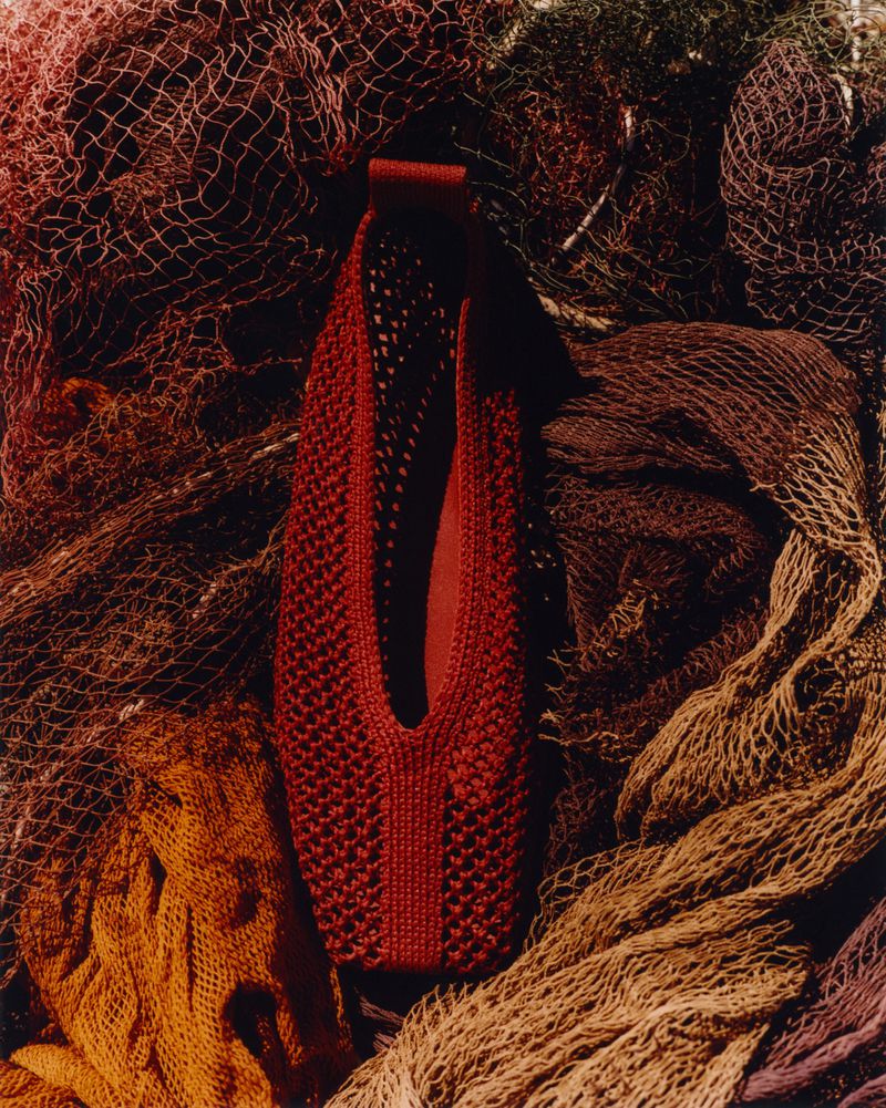Red mesh bag on multicolored netting. Knitted bag with handle sits atop piled fishing nets in red, brown, yellow, and tan hues.