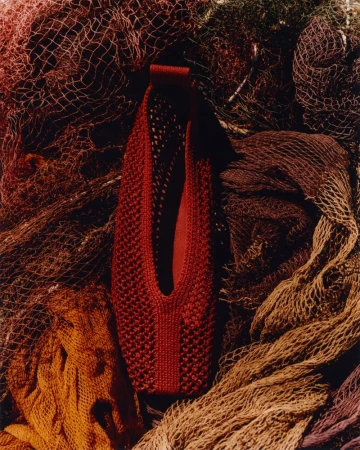 Red mesh bag on multicolored netting. Knitted bag with handle sits atop piled fishing nets in red, brown, yellow, and tan hues.