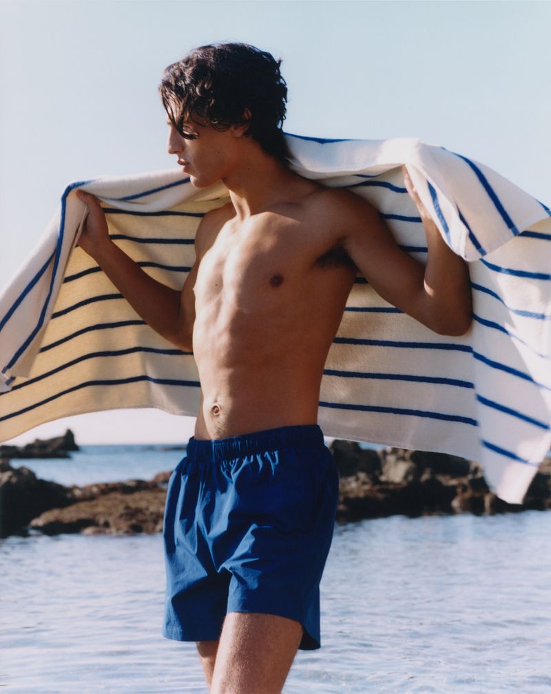 Young man on beach with blue swim trunks and striped towel draped over shoulders, clear sky and water, relaxed summertime vibes.