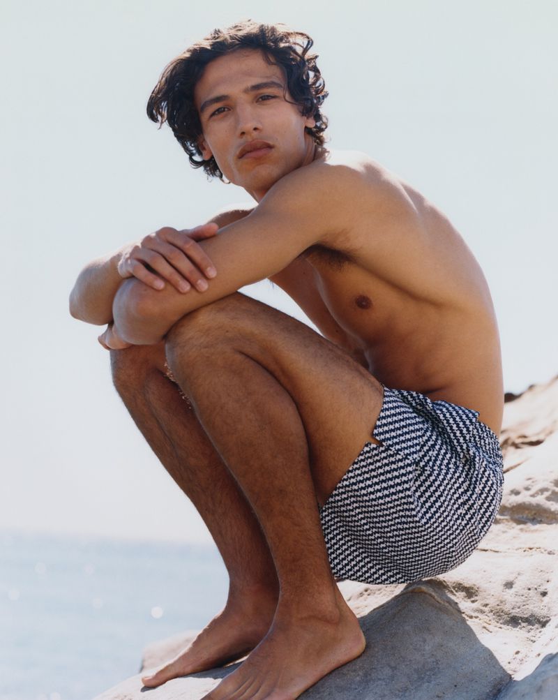 Shirtless man with dark hair poses on a rock formation wearing patterned swim trunks, with an ocean backdrop under a bright sky.