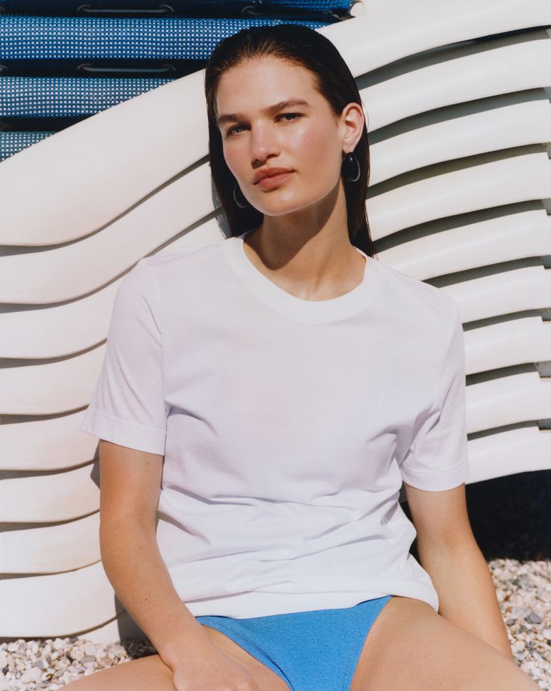Adult woman poses in a white t-shirt and blue bikini bottoms, seated on a beach with stacked surfboards in the background.
