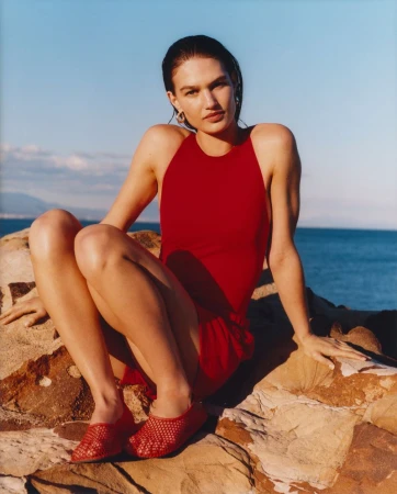 A woman in a red swimsuit and matching shoes poses on rocks by the sea under a bright blue sky in a sunny outdoor fashion shot.