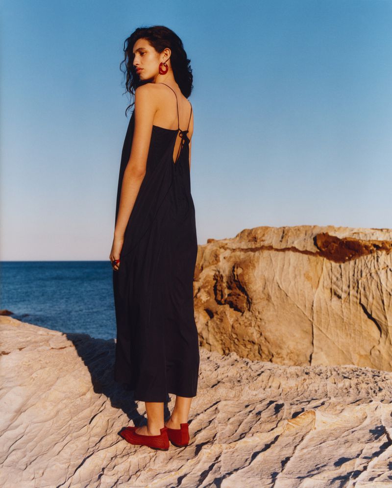 Woman in a flowing black dress stands on a rock formation by the sea, wearing red earrings and shoes, captured in an eye-level medium shot.