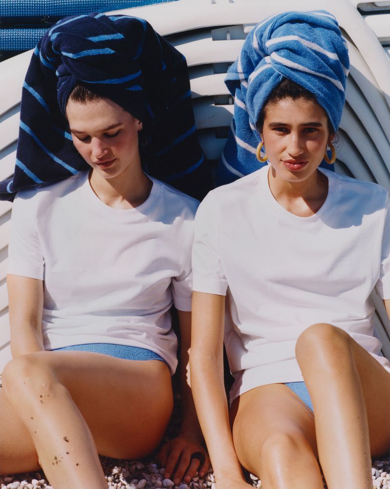 Two women with towels wrapped on their heads, wearing white t-shirts and blue bottoms, are sitting close together on a pebble beach.