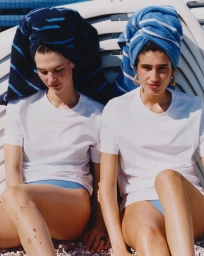 Two female models wearing white t-shirts and striped towels on their heads seated against white loungers.