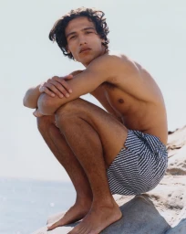 Shirtless man wearing patterned swim shorts squatting on a rocky shore against a clear sky