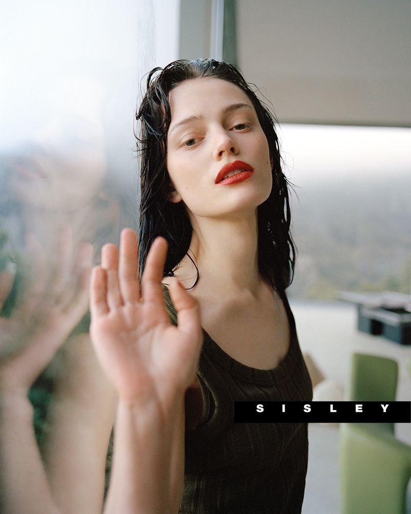 Portrait of a young woman with wet dark hair, red lipstick and wearing a dark top, standing near a glass wall.