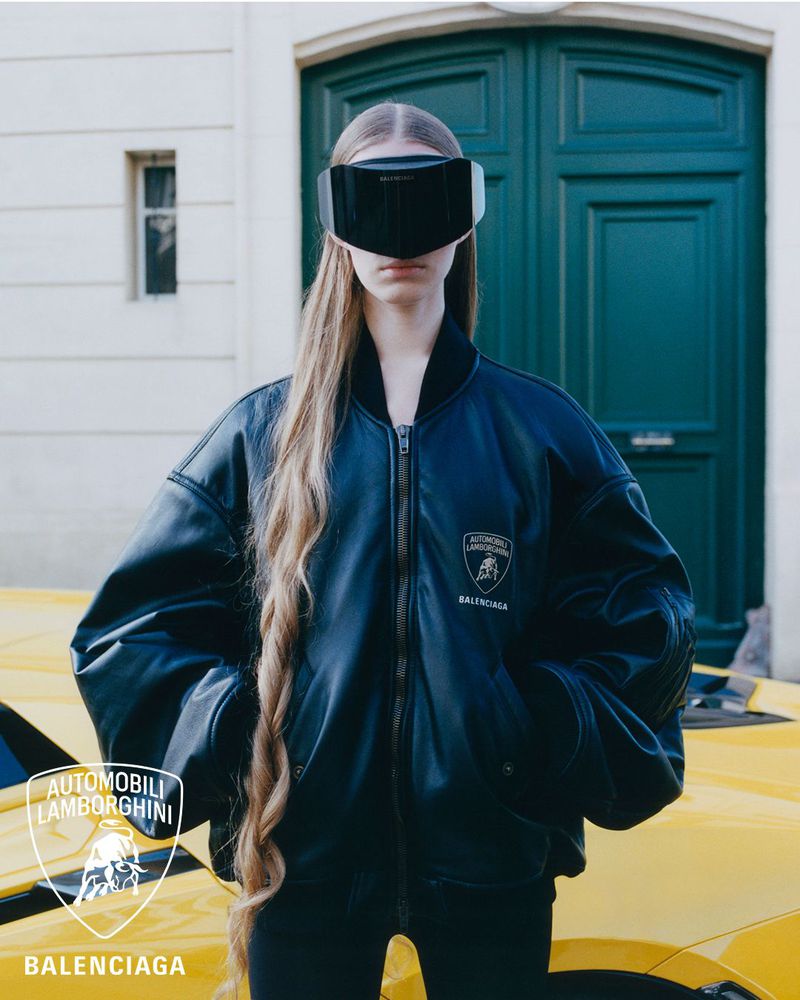 Model in visor and black Balenciaga Lamborghini bomber jacket poses with a yellow Lamborghini. Fashion photography.