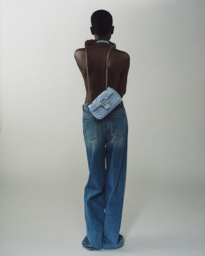 A person is facing away from the camera wearing jeans and a light blue purse with a silver chain. The background is a solid off-white.
