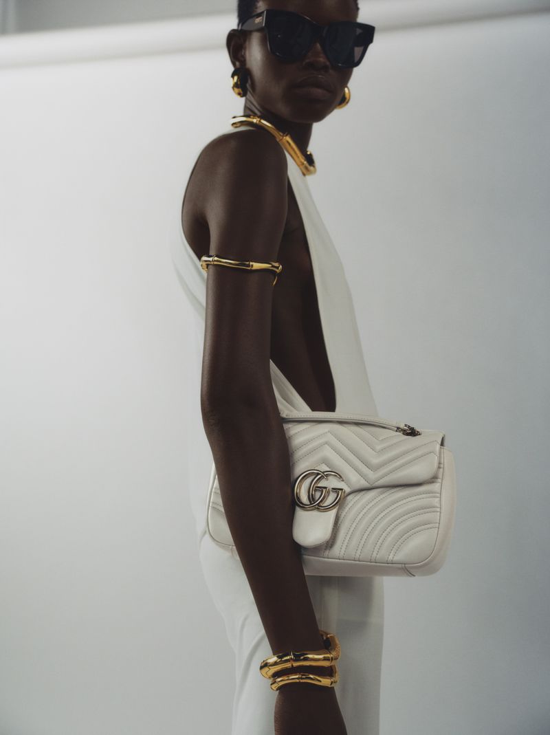 Fashion shot of a Black woman wearing a white dress, sunglasses, and gold jewelry holding a Gucci bag against a white background.