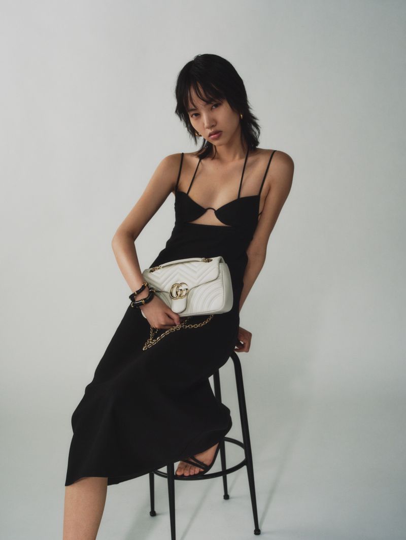 Elegant Asian woman in a black dress sitting on a stool with a white Gucci purse and gold chain, studio shot on a light background.