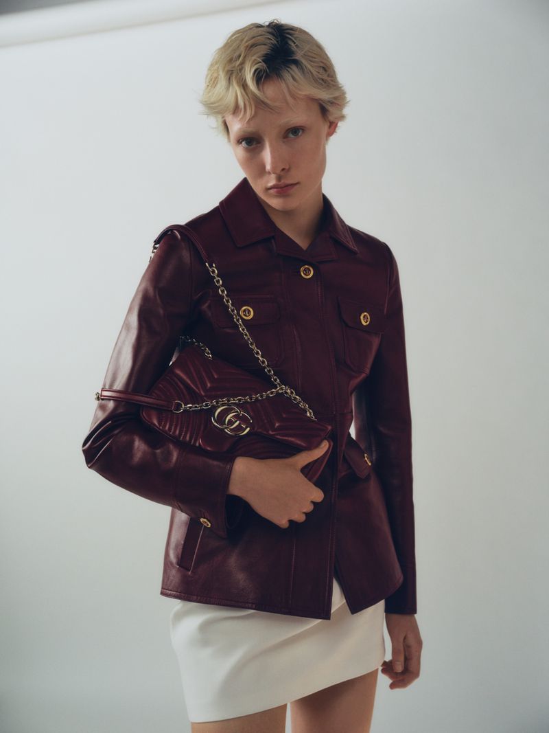A woman in a red leather jacket holds a Gucci Marmont handbag against a neutral backdrop, showcasing a high-fashion editorial shot.