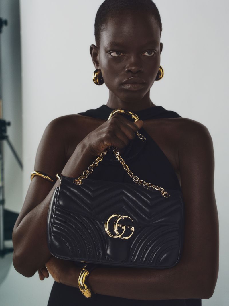 Close-up fashion portrait of a woman with dark skin and short hair holding a black Gucci purse with gold jewelry in a studio.