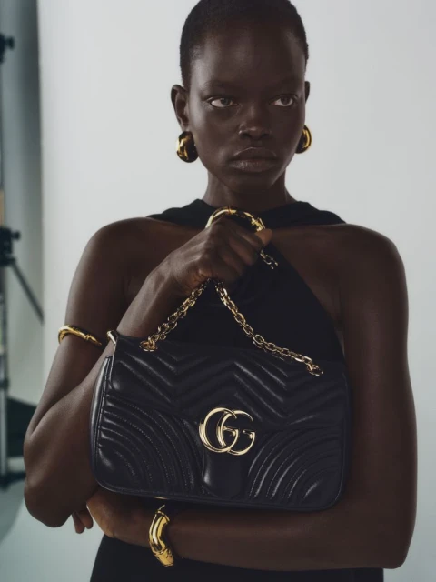 Close-up fashion portrait of a woman with dark skin and short hair holding a black Gucci purse with gold jewelry in a studio.