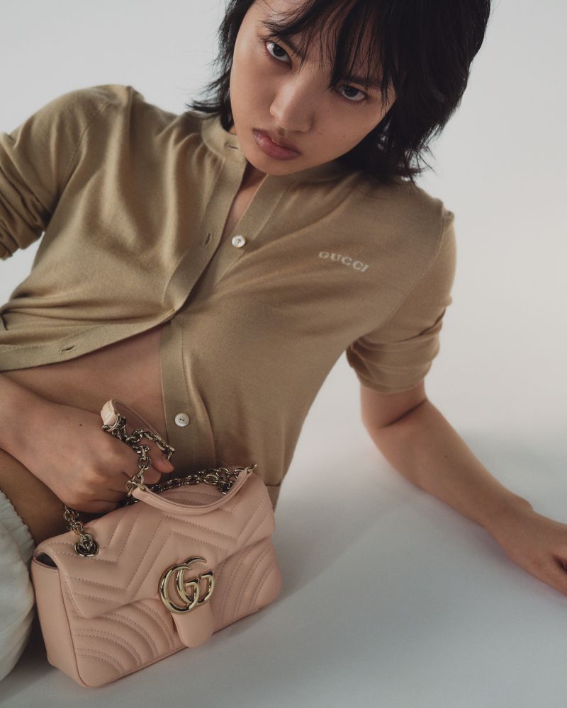 Portrait of a young Asian woman in a beige cardigan with a Gucci purse, posing against a white backdrop in a studio setting.