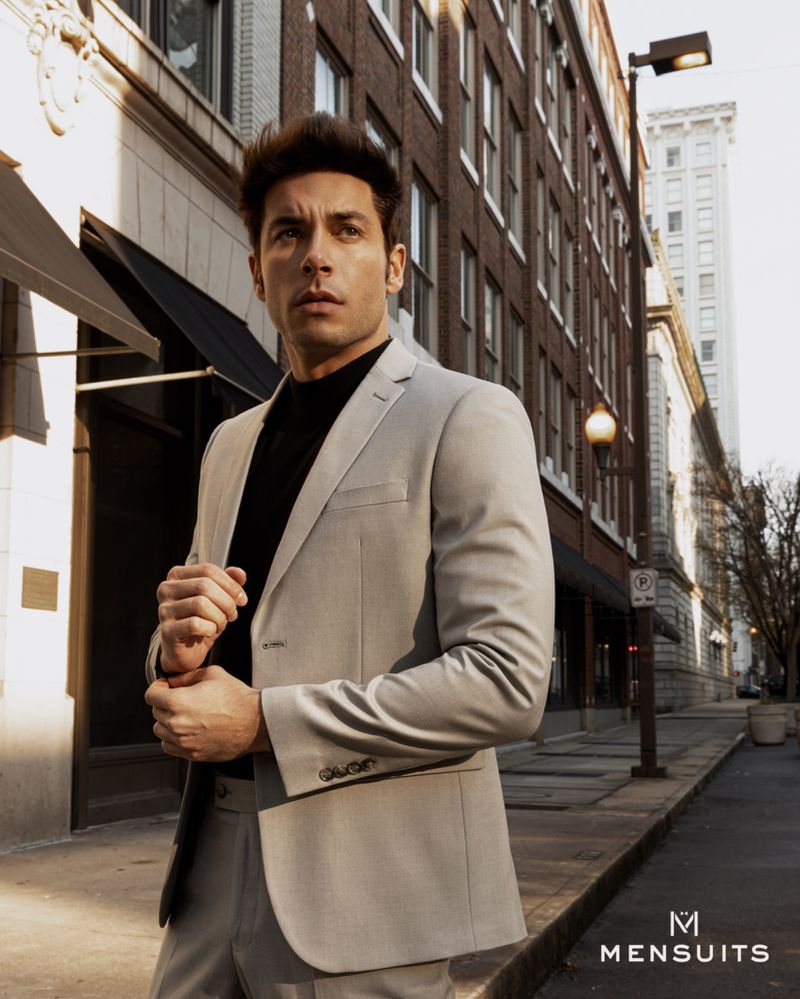 Man in light grey suit poses on a city street with brick buildings, looking off-camera in an outdoor fashion style shoot. 