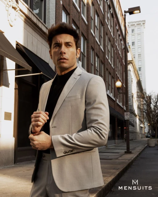 Man in light grey suit poses on a city street with brick buildings, looking off-camera in an outdoor fashion style shoot. 